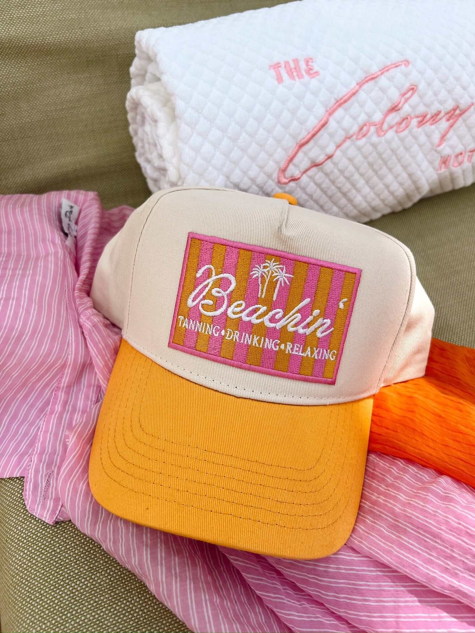 Cream and yellow trucker hat with colorful pink and gold "Beachin' Tanning Drinking Relaxing" patch on front, styled on pink and orange fabrics.