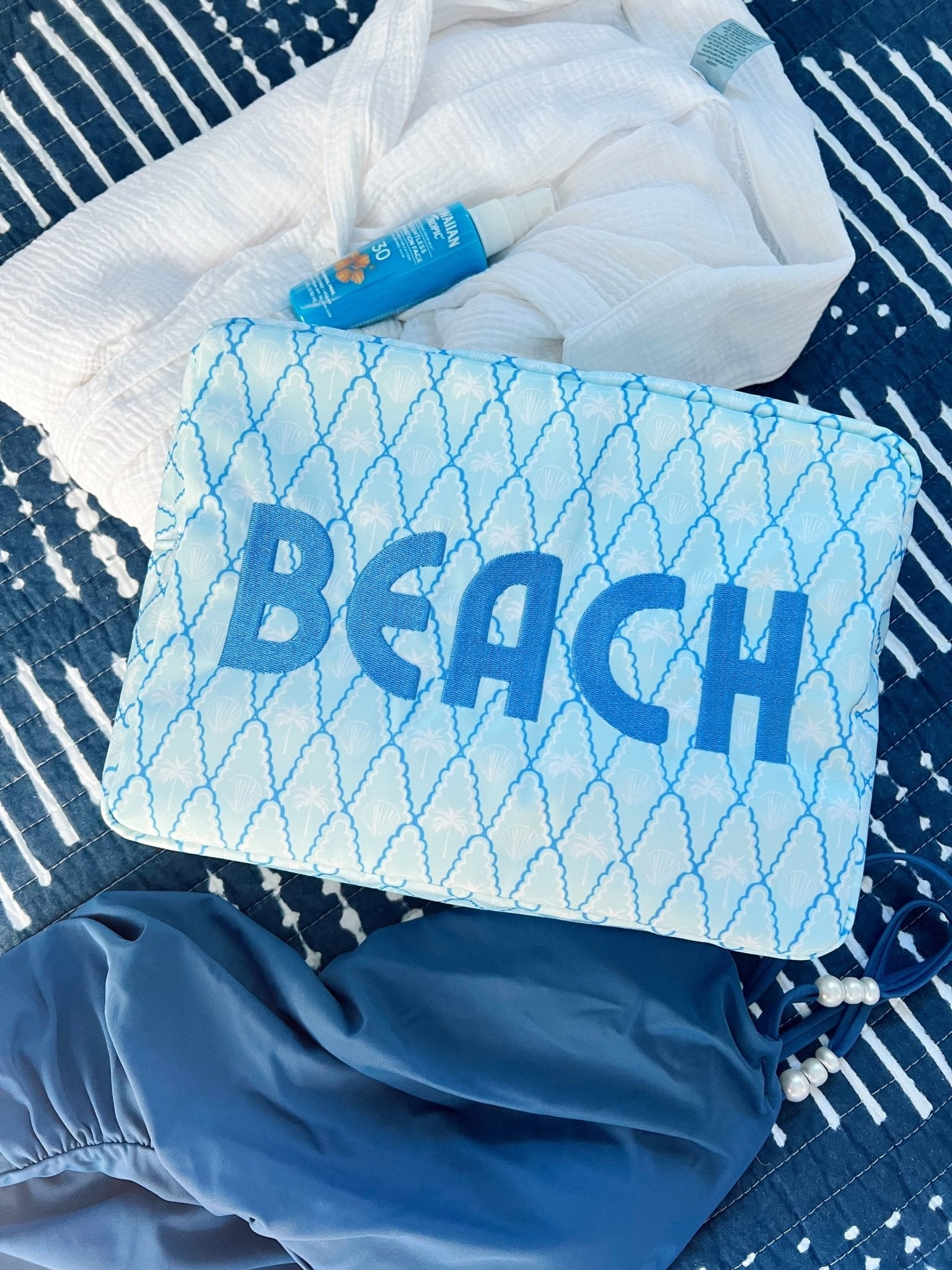 Blue and white mesh makeup bag with "BEACH" text, displayed with sunscreen and beach towel on striped fabric.