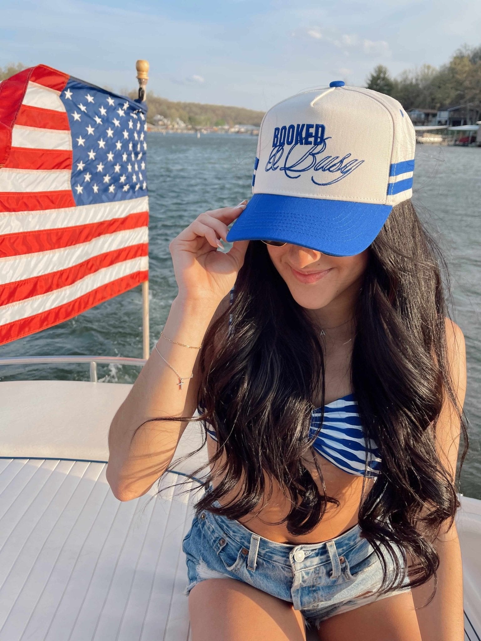 Royal blue and cream vintage trucker hat with "Booked and Busy" script text, styled on boat with American flag background.