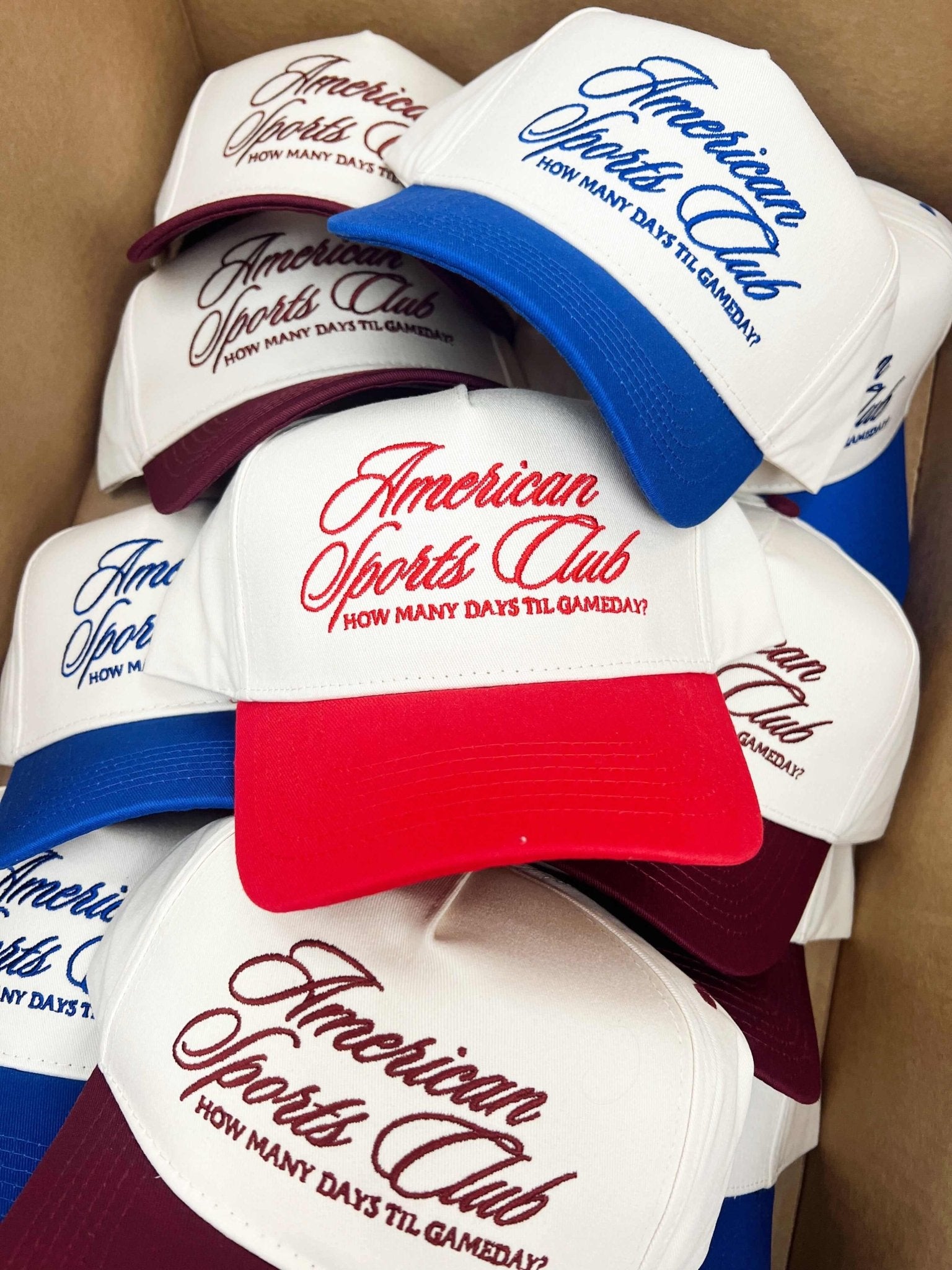 Stack of vintage trucker hats with red, white, and blue colorways featuring American Sports Club script logo and "How Many Days Till Gameday" text.