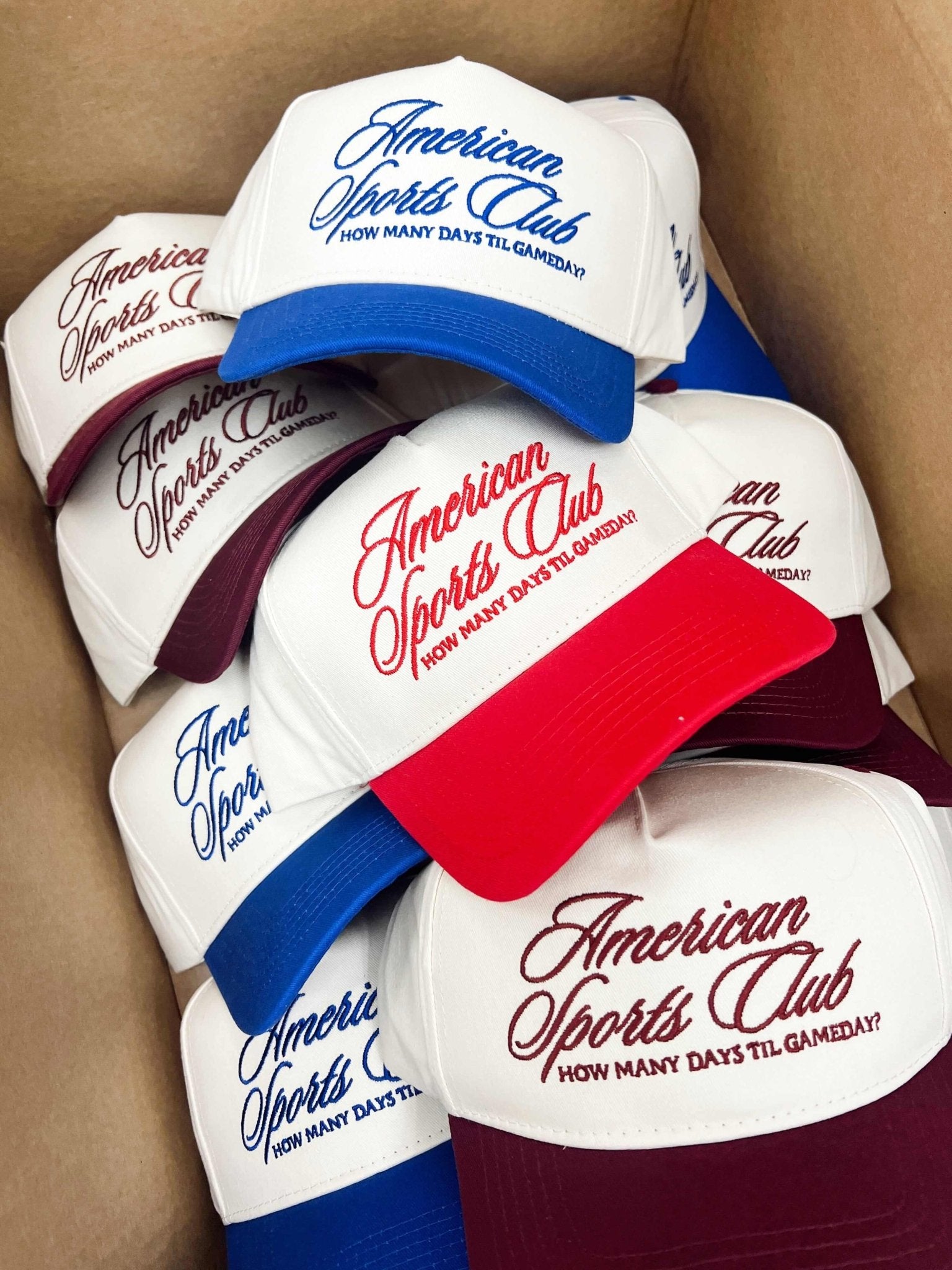 Stack of vintage trucker hats with "American Sports Club" script text in blue, red, and burgundy on white panels with matching colored mesh backs.