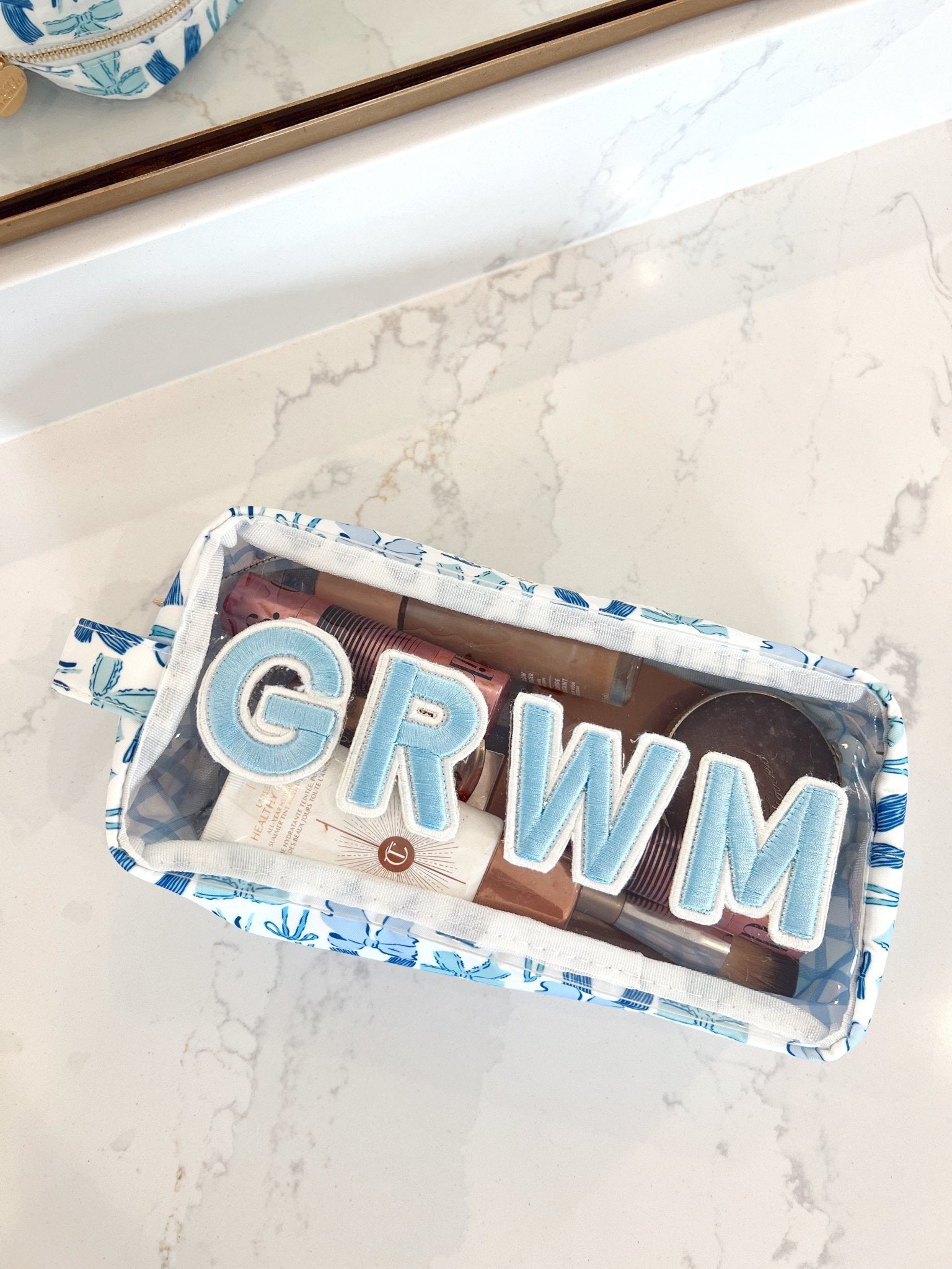 Clear makeup bag with blue bow pattern trim and white "GRWM" lettering, filled with cosmetics and brushes.