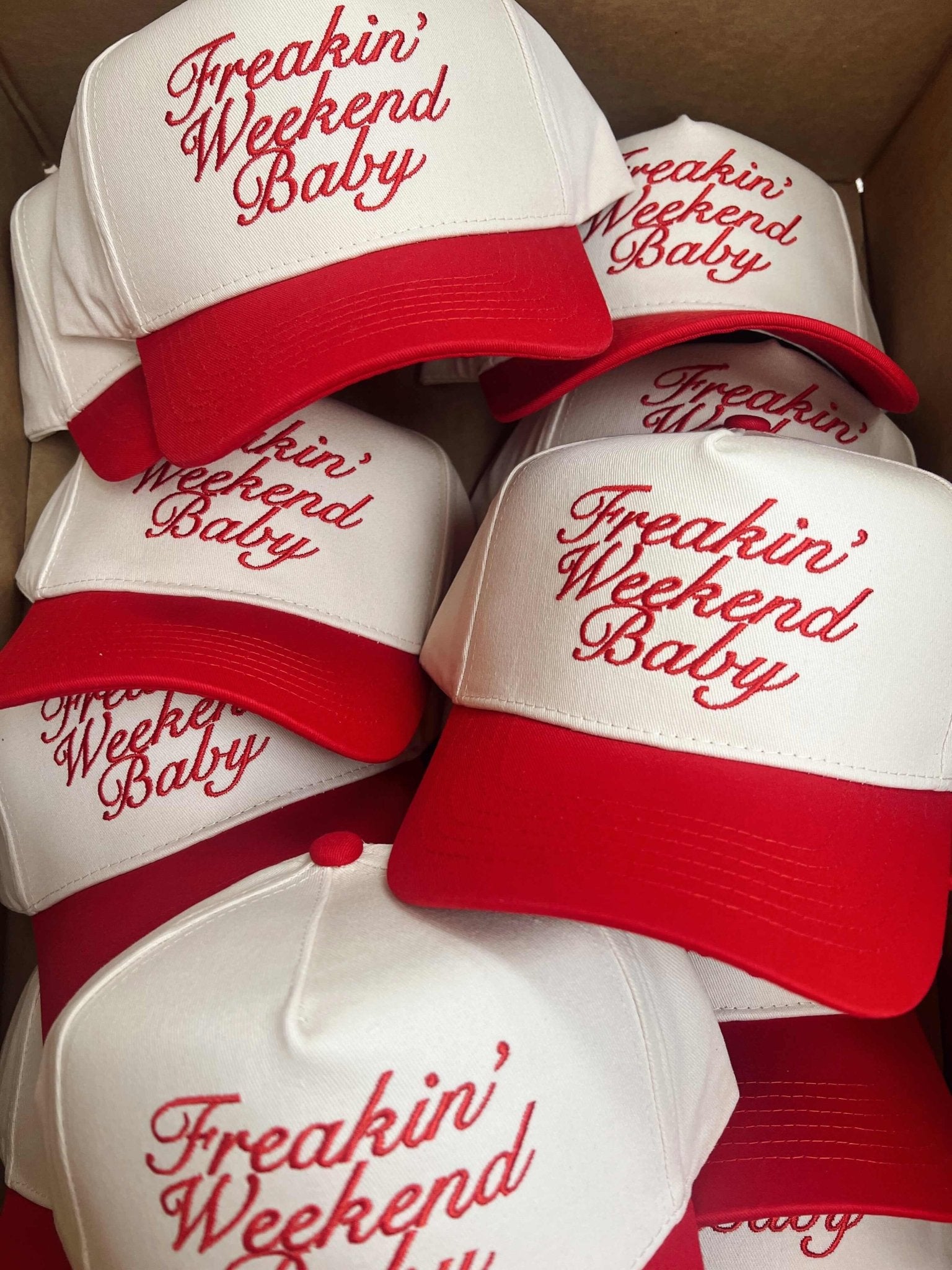 White trucker hats with red "Freakin' Weekend Baby" embroidery and red visor straps stacked together.