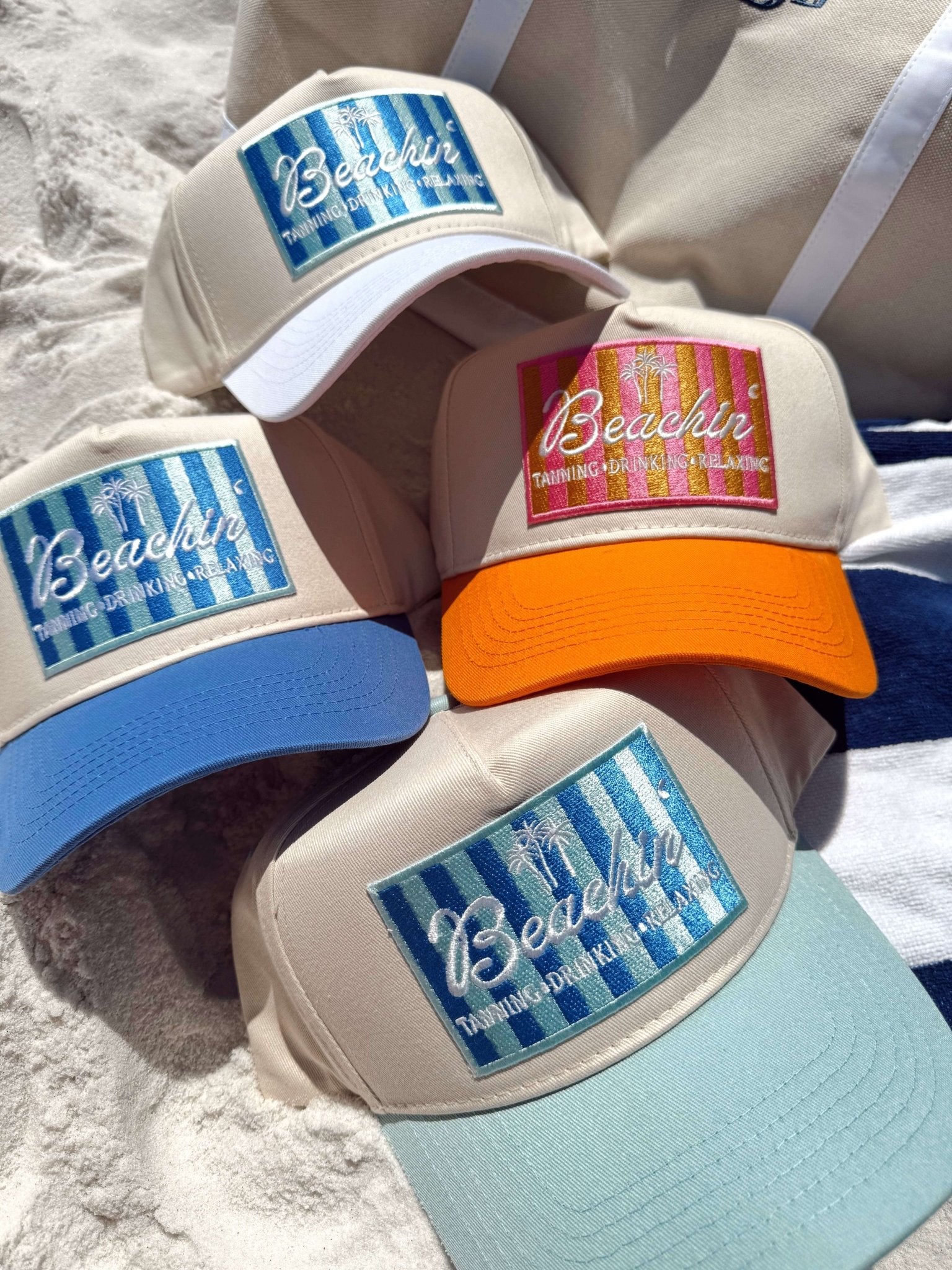 Collection of vintage trucker hats with "Beachin'" designs in blue striped, orange, and light blue colorways on sand.