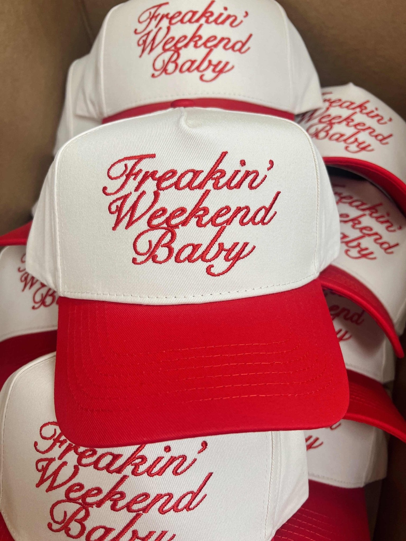 White and red trucker hats with "Freakin' Weekend Baby" embroidered in red script across the crown.