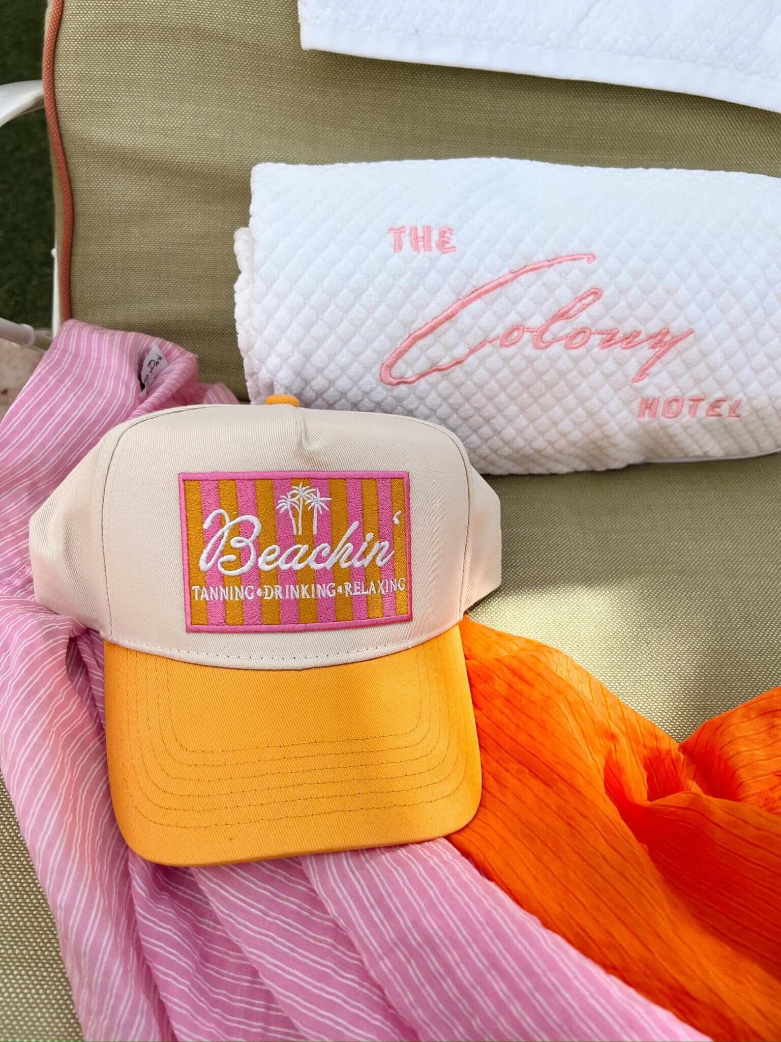 Cream and yellow trucker hat with colorful "Beachin'" patch, styled with beach pillows and bright fabrics outdoors.