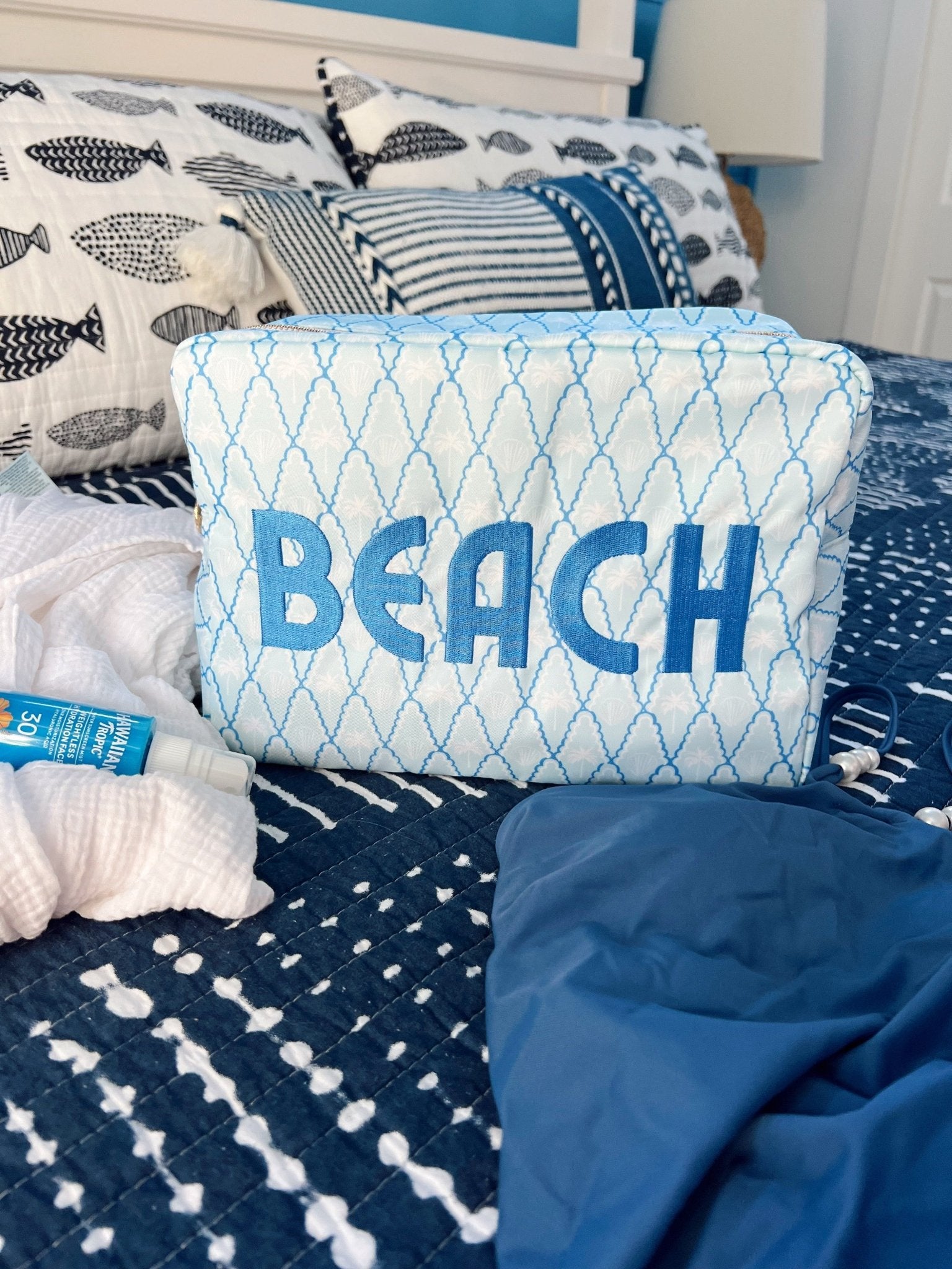 White quilted cosmetic bag with blue diamond pattern and "BEACH" text, styled on bedding