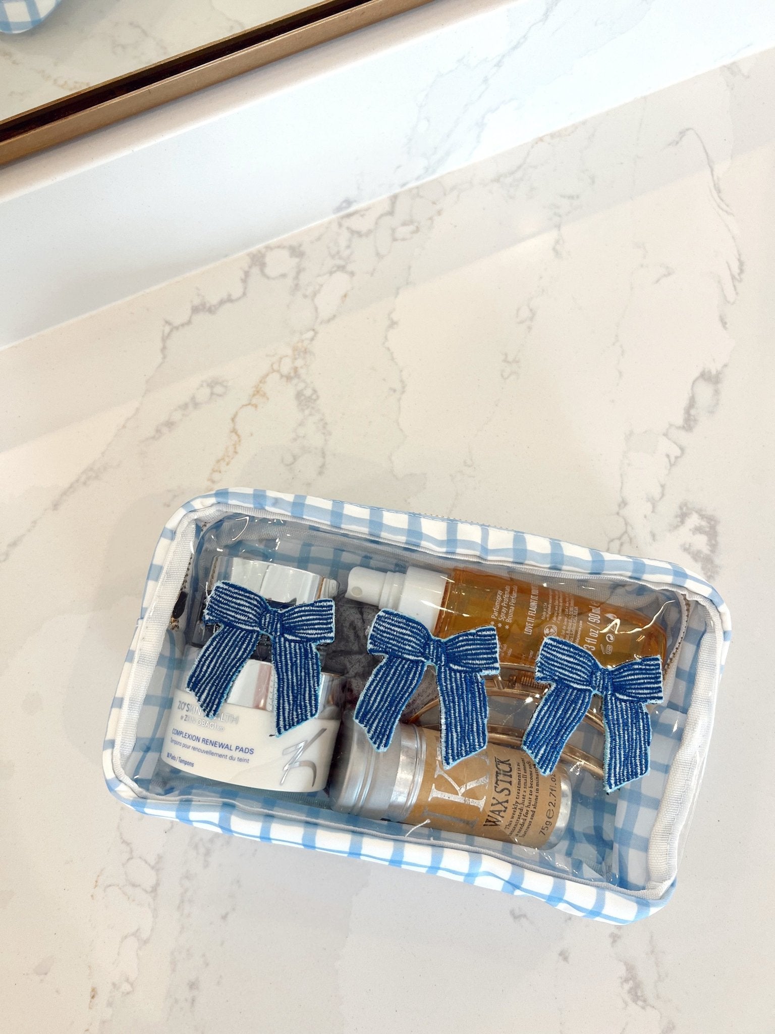 Clear plastic makeup bag with blue and white gingham trim containing three decorative bows in blue striped and mustard patterns.