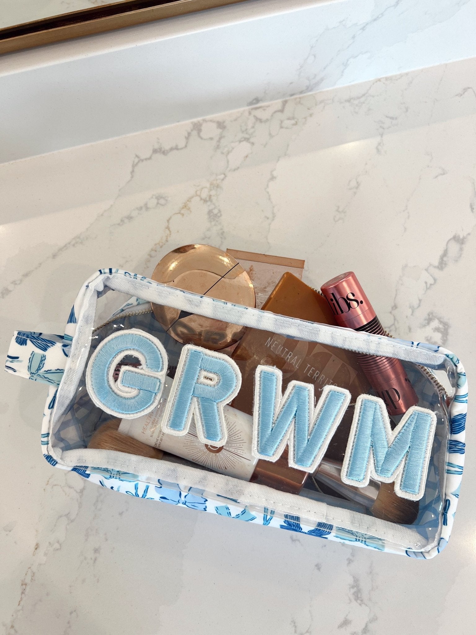 Clear makeup bag with blue bow pattern trim and blue "GRWM" lettering, filled with cosmetics and accessories on marble surface.