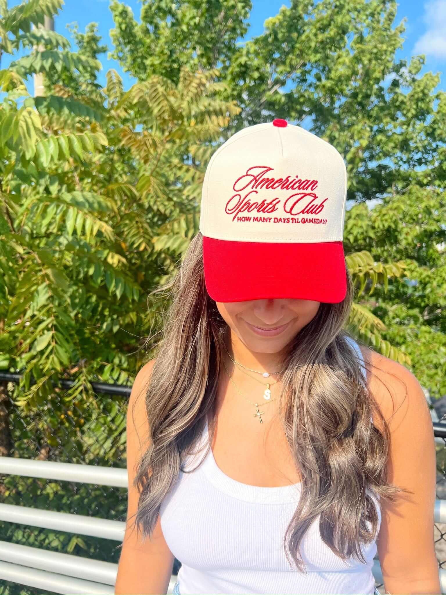 Red and cream trucker hat with "American Sports Club" embroidered text and curved brim, worn outdoors.
