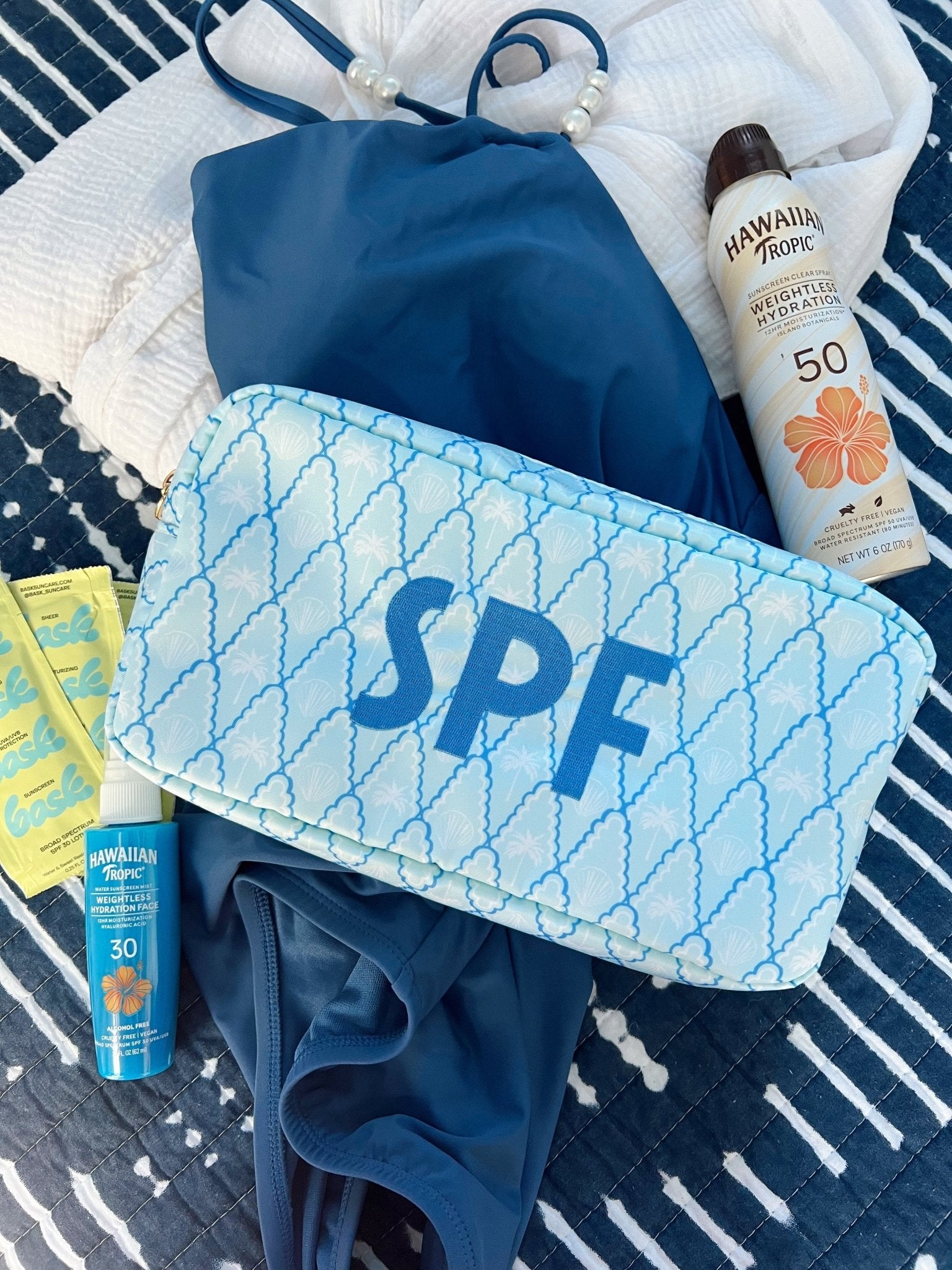 Blue and white quilted makeup bag with SPF text, surrounded by sunscreen bottles and beach accessories on striped towel.