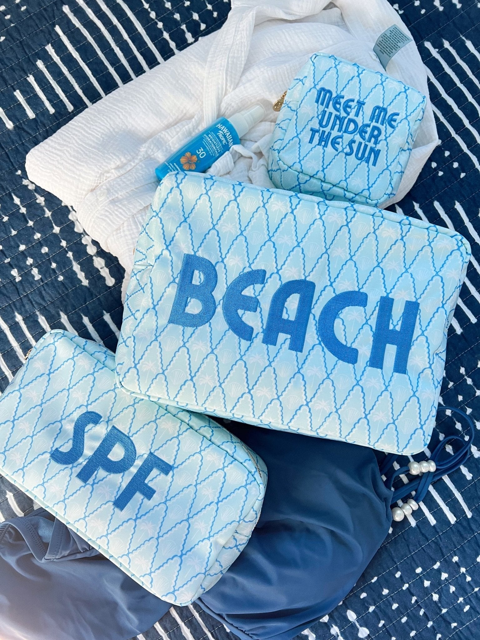 Blue fishing net patterned makeup bags with "BEACH" and "SPF" text, displayed with sunscreen bottle on striped beach towel.