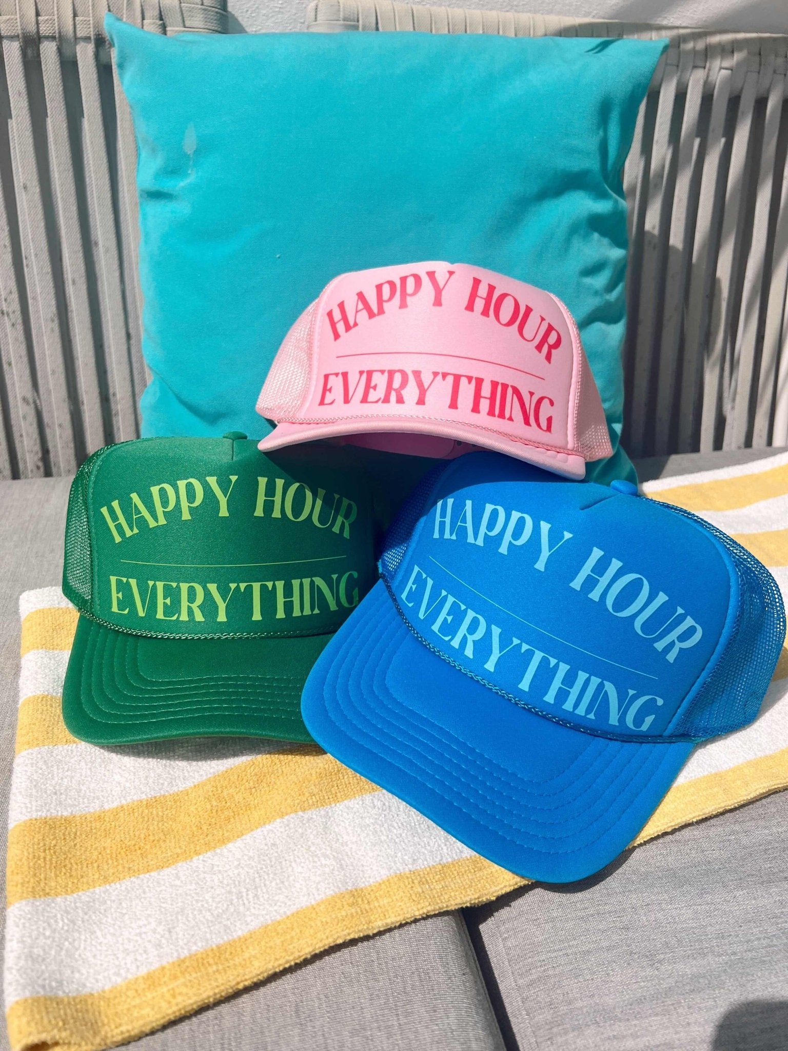 Three trucker hats in pink, green, and blue with "Happy Hour Everything" text, displayed on a turquoise pillow and striped towel.