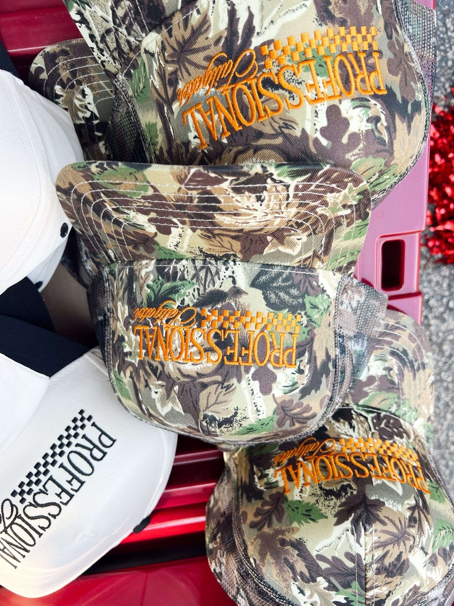 Vintage camo trucker hat with orange embroidered text and mesh back panels, stacked with white cap below