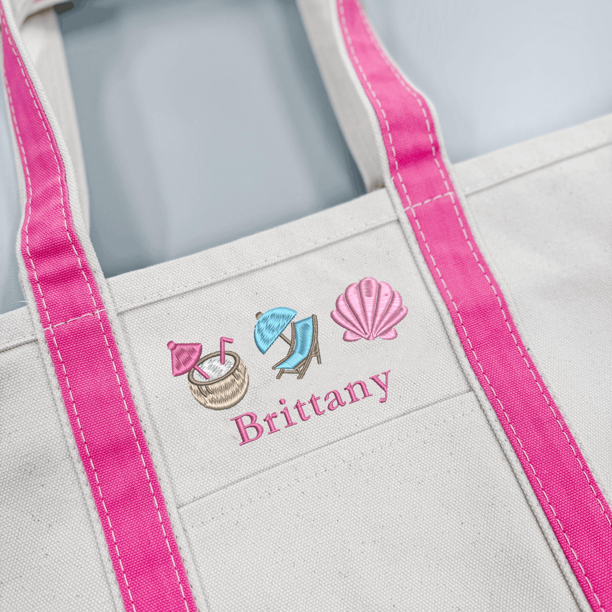 Close-up of pink embroidery on a personalized canvas tote featuring the name Brittany with coconut drink, beach chair, and seashell icons.
