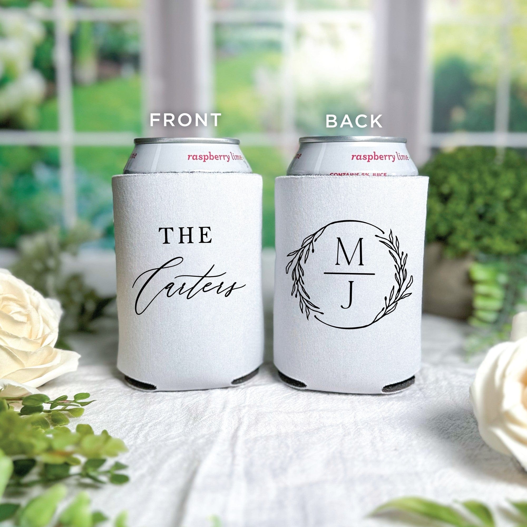 Two white neoprene can coolers are personalized for a wedding, one featuring a custom calligraphic text design and the other an elegant monogram encircled by a delicate leaf wreath.