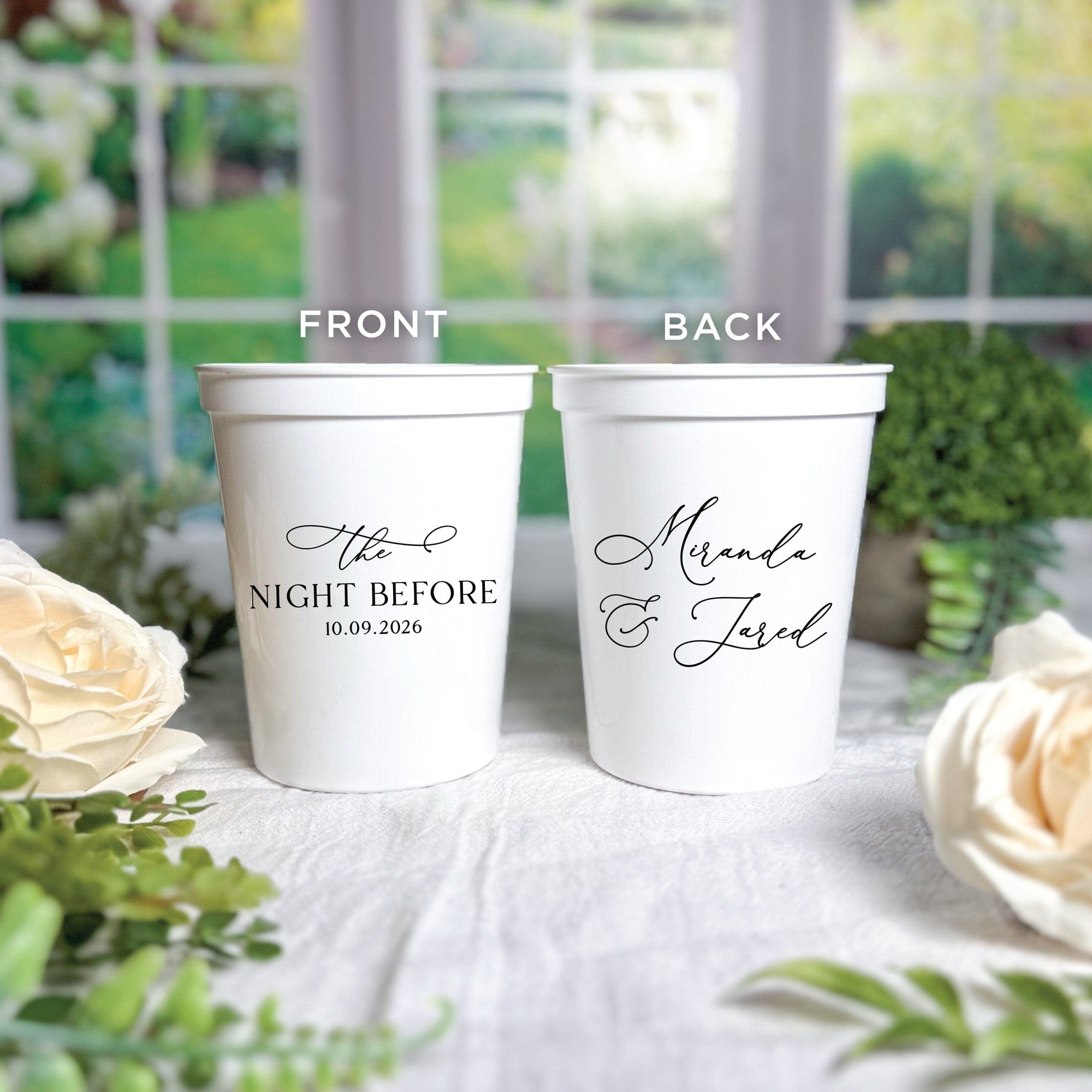 Two personalized white plastic wedding party cups feature custom black script text on both the front and back, presented on a white tablecloth with green foliage and white roses.