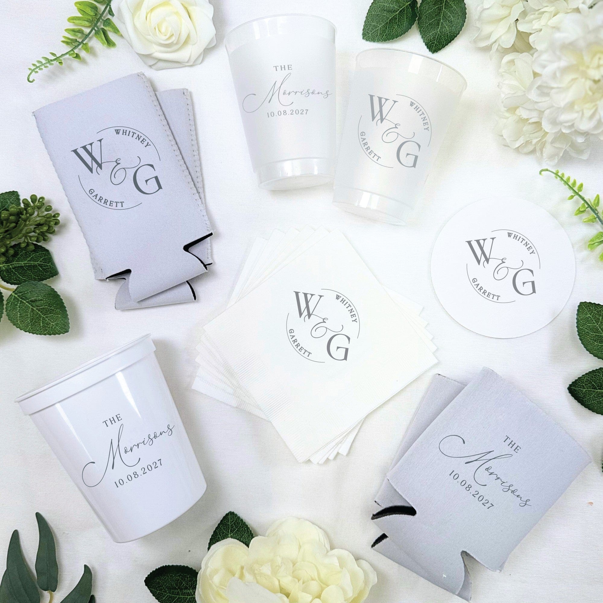 A collection of personalized wedding party supplies including translucent plastic cups, white paper napkins, white paper coasters, and light grey koozies, all custom-printed for a celebratory event.