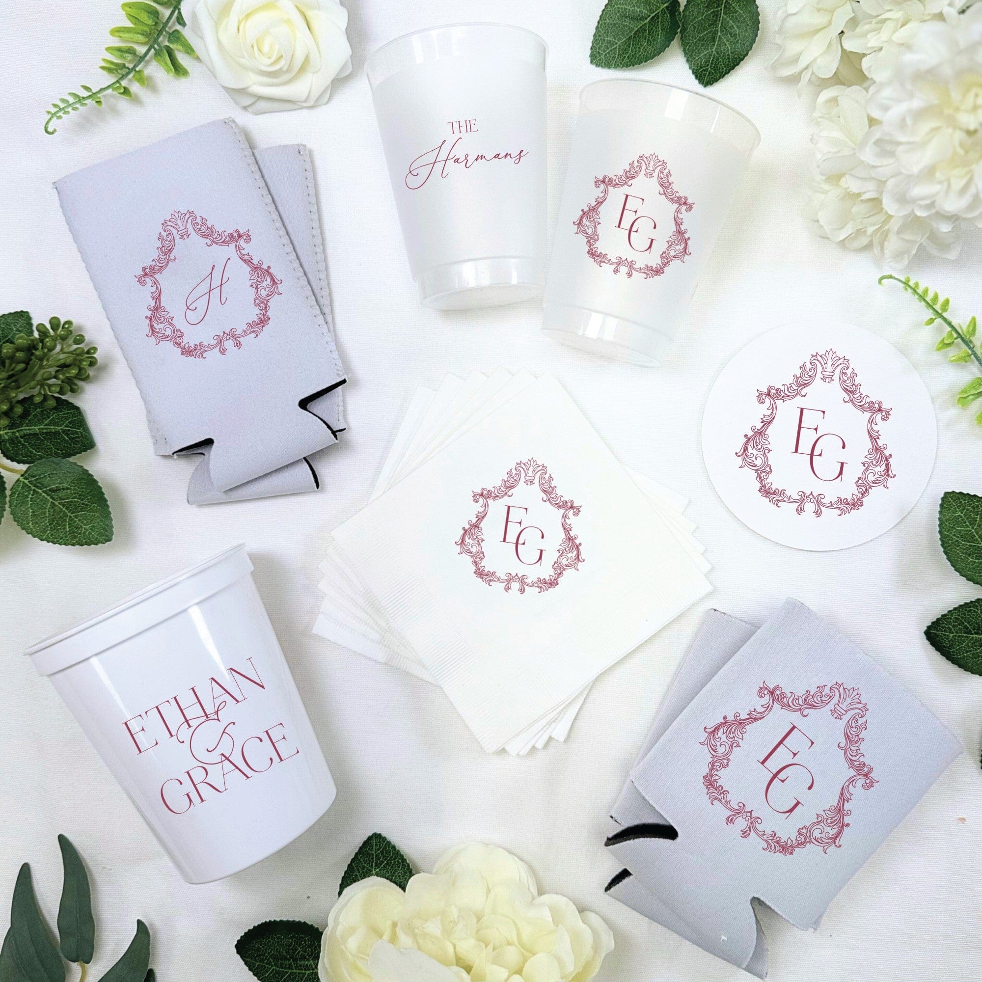 This wedding decor bundle features frosted and white plastic cups, light grey neoprene can coolers, white paper napkins, and round paper coasters, all adorned with a matching elegant decorative design in a deep red color, suitable for a wedding or special event.