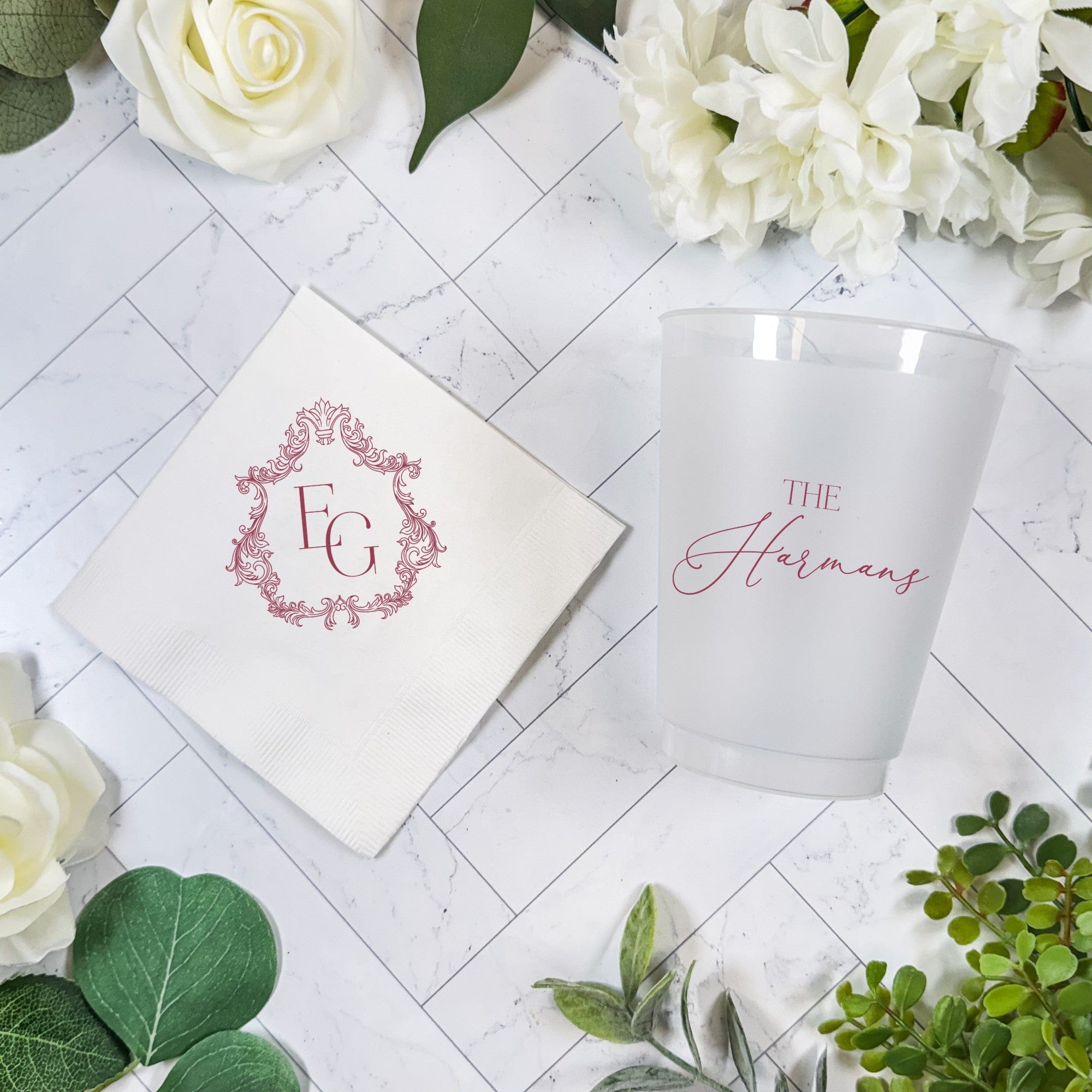 A personalized wedding bundle featuring a white paper napkin with an ornate monogram design and a frosted plastic cup with elegant custom script, perfect for reception decor.