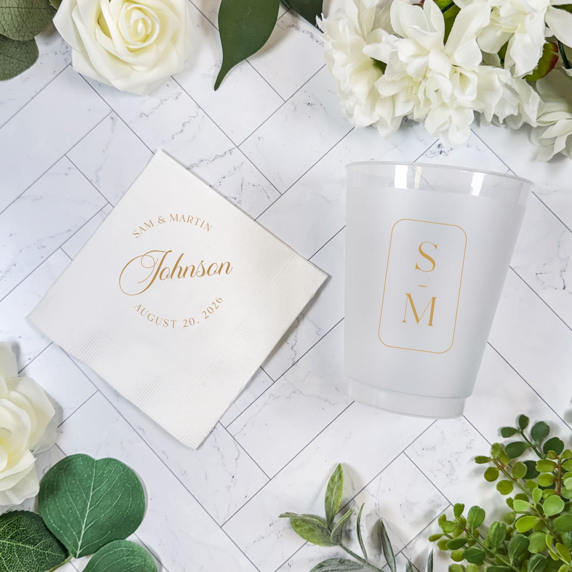 A personalized wedding bundle featuring a white paper napkin with gold foil text and a frosted plastic cup with a gold foil initial design, set on a marble background with white flowers and green leaves.
