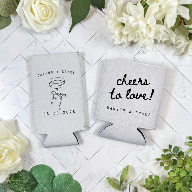 Two light grey fabric wedding can coolers are displayed, one featuring a champagne coupe illustration with a ribbon detail and the other a decorative script phrase above a line of block text, both serving as personalized drink insulators.