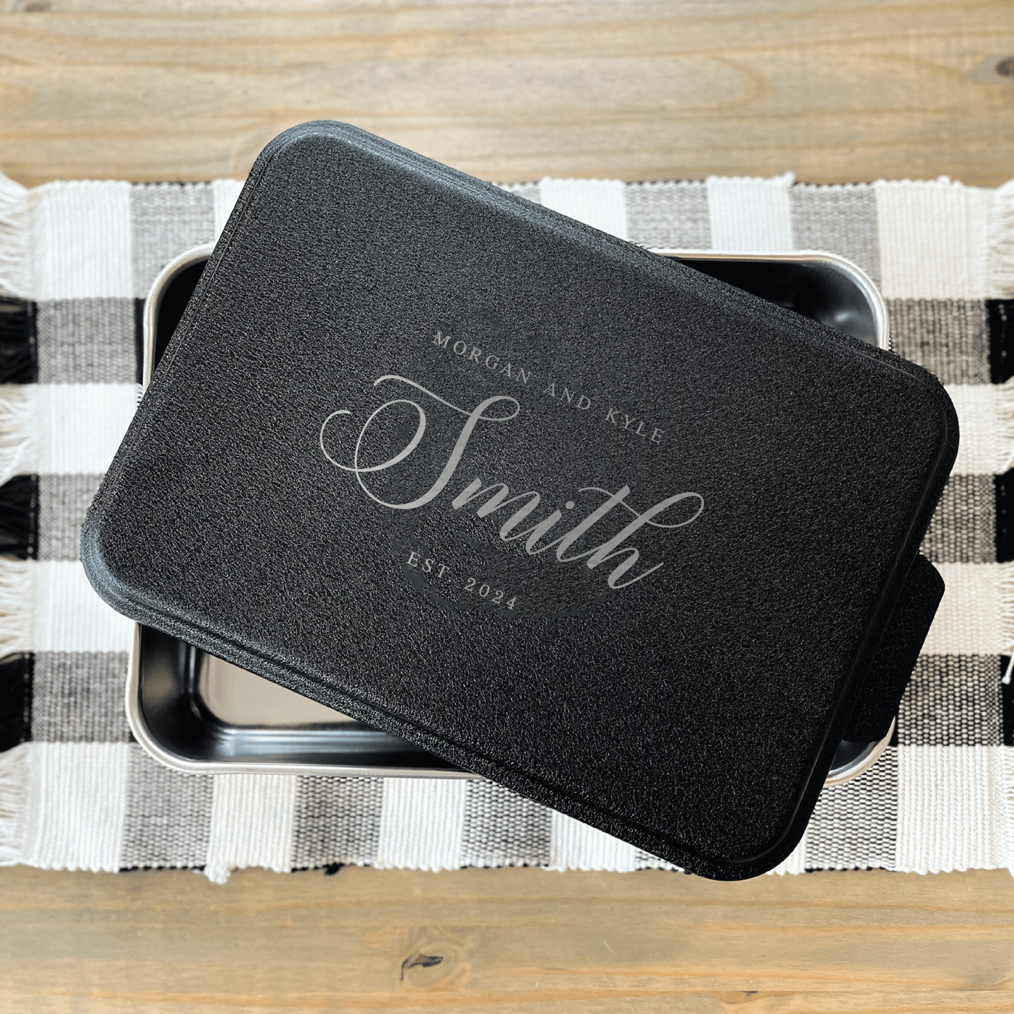 Black personalized cake pan with "Smith" engraved in script, displayed open on a checkered kitchen towel.