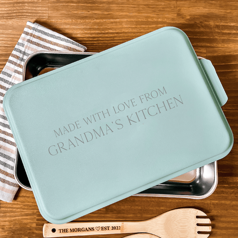 Light blue cake pan with "Made With Love From Grandma's Kitchen" engraved on lid, displayed with wooden utensils and striped cloth on wood surface.
