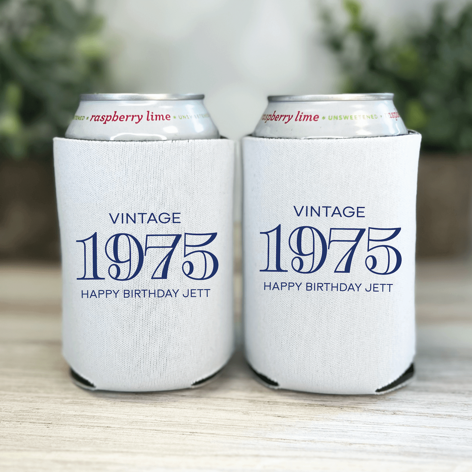 Two white can coolers with "Vintage 1975" and "Happy Birthday Jett" personalized text in navy blue, holding canned beverages outdoors.