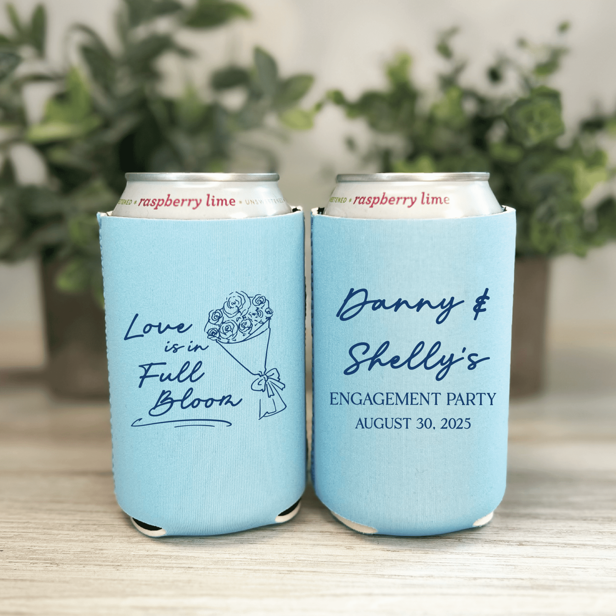 Two light blue can coolers with personalized text, one reading "Love Is In Full Bloom" with floral design, the other "Danny & Shelly's Engagement Party August 30, 2025"