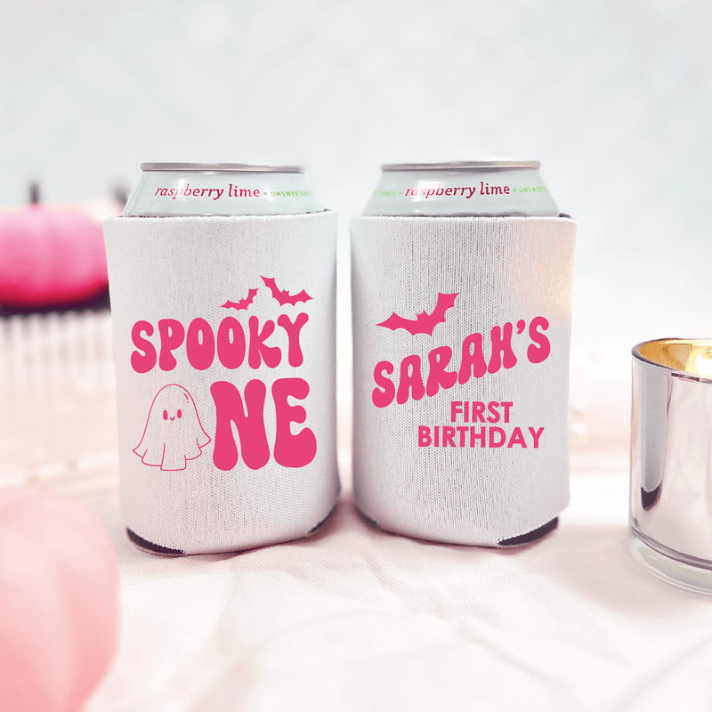 Two white can coolers with hot pink text and designs; left reads "SPOOKY ONE" with ghost graphic, right reads "SARAH'S FIRST BIRTHDAY" with bat design.