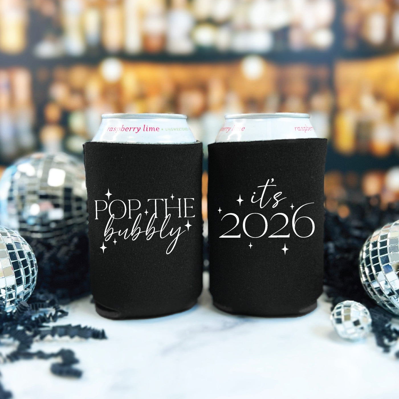Two black fabric can coolers featuring white celebratory designs are shown keeping slim cans cold amidst disco balls and black festive decor, perfect for a New Year's Eve party.
