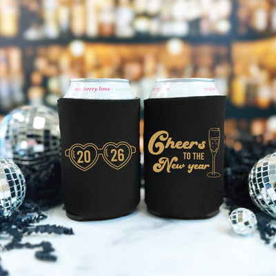 Two black fabric can coolers, each featuring a metallic gold design, display festive heart-shaped glasses on one and a champagne flute on the other, ideal for keeping beverages cold at a New Year's Eve celebration.