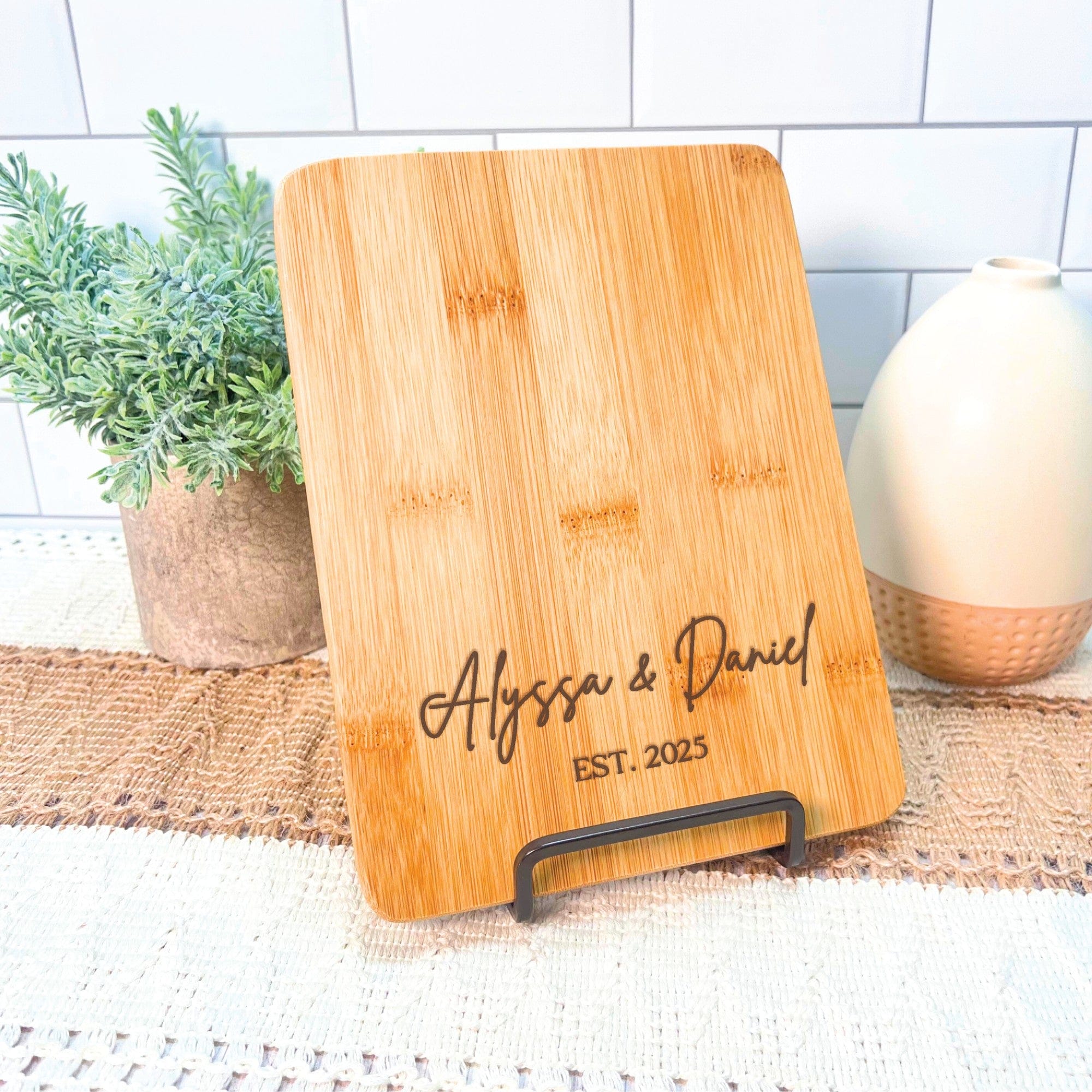 A rectangular bamboo cutting board with rounded corners, featuring custom laser-engraved text, displayed on a black metal stand, ideal for wedding or engagement decor.