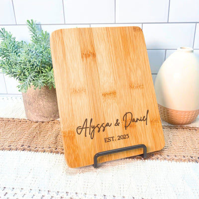 A rectangular bamboo cutting board with rounded corners, featuring custom laser-engraved text, displayed on a black metal stand, ideal for wedding or engagement decor.