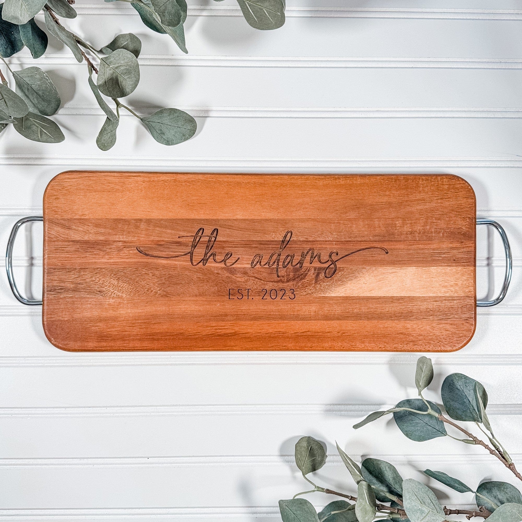 A rectangular acacia wood charcuterie board with rounded corners, featuring engraved personalization and two silver metal handles, suitable as a wedding or housewarming gift.