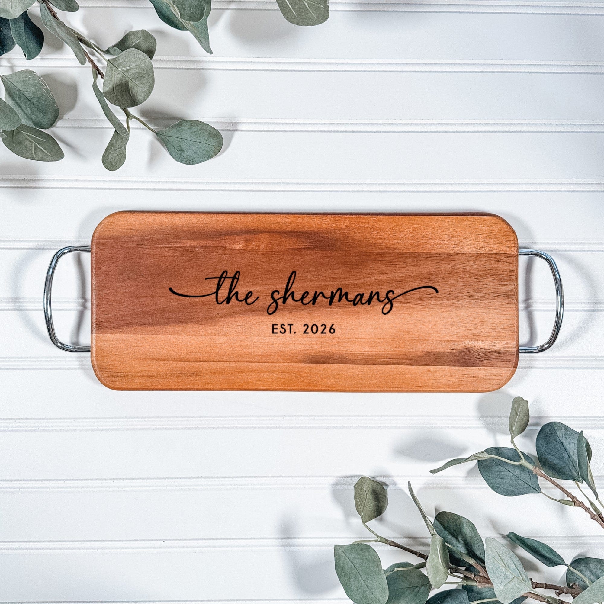 A rectangular acacia wood serving board with silver metal handles, custom engraved with a design, ideal for a wedding or housewarming gift.