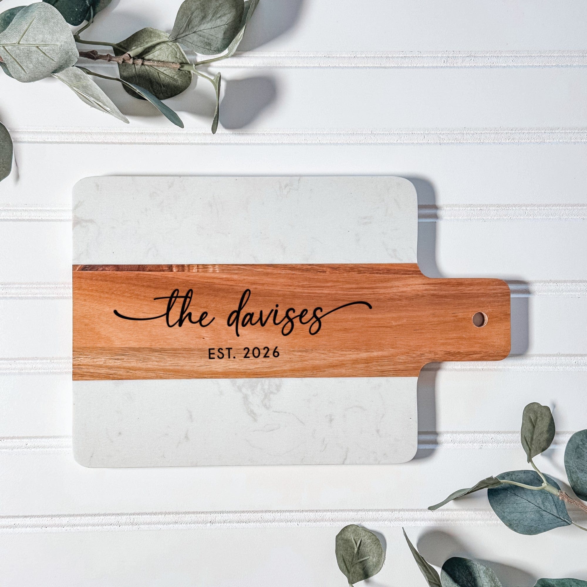 A personalized two-toned rectangular charcuterie board featuring a white marble top and a natural wood handle, ideal as a wedding or housewarming gift.