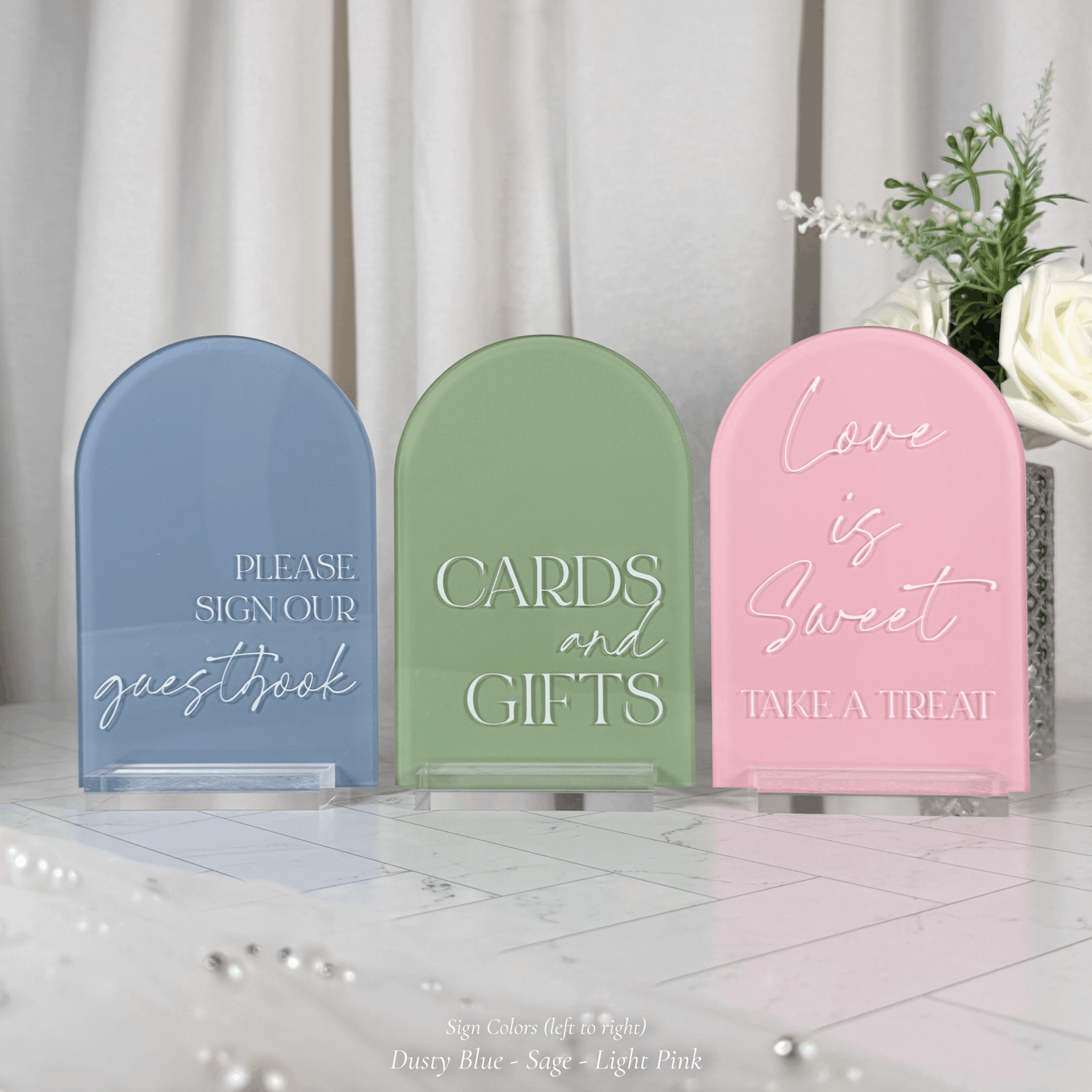 Three arched acrylic wedding signs in dusty blue, sage green, and light pink on clear stands with a flowing curtain backdrop and fresh florals, reading Please Sign Our Guestbook, Cards and Gifts, and Love is Sweet Take a Treat.