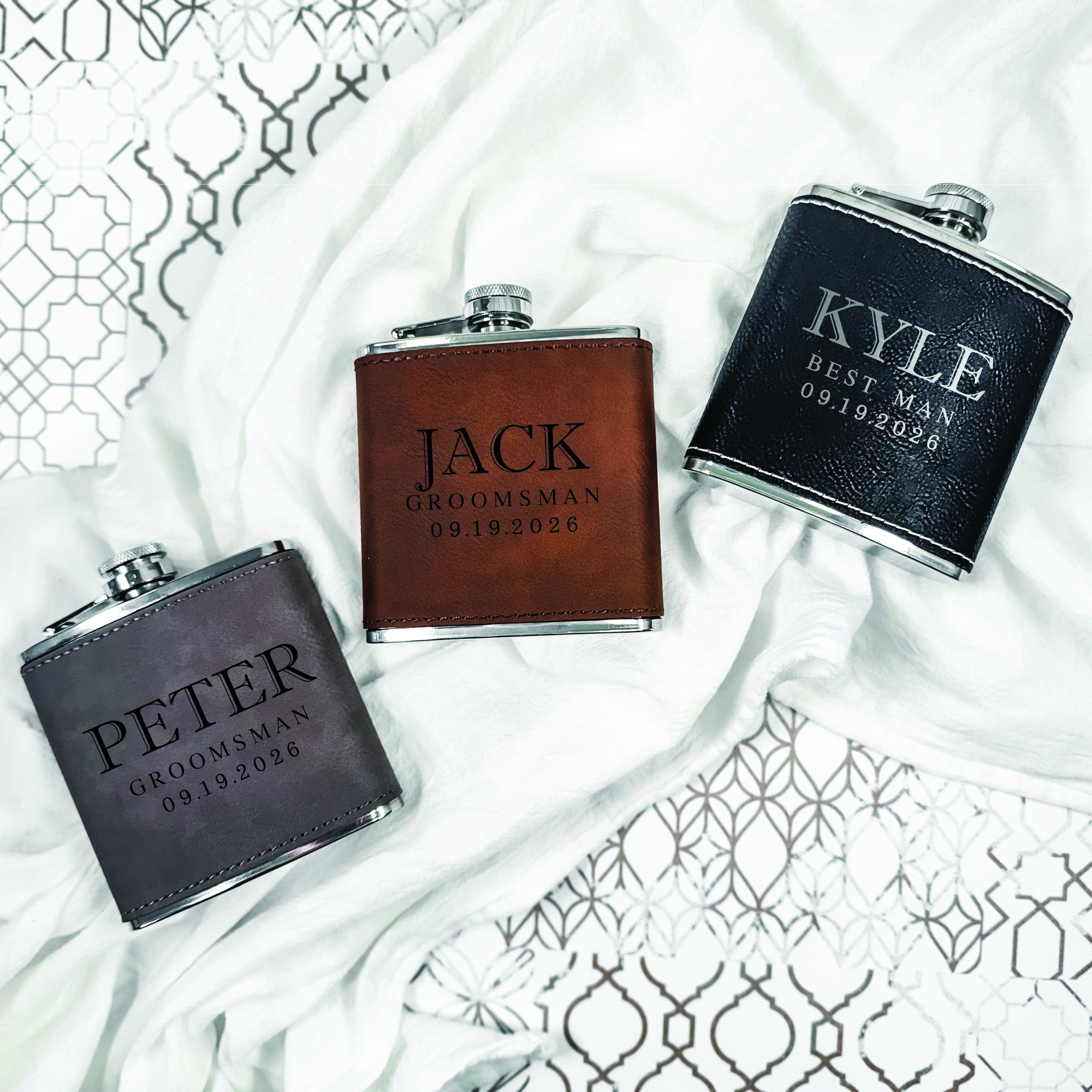 Three personalized stainless steel hip flasks featuring faux leather wraps in brown, black, and gray, suitable as groomsmen gifts for a wedding.