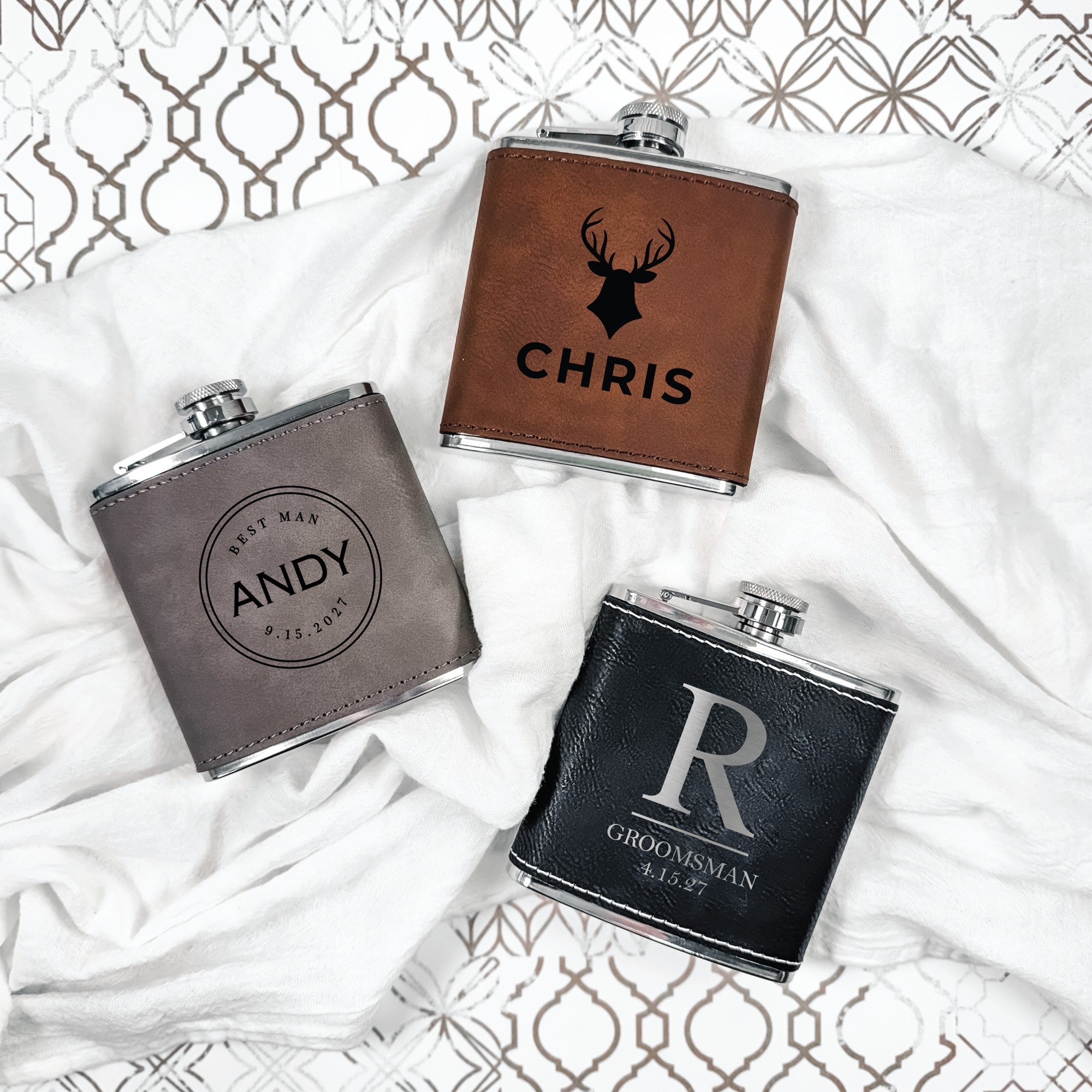 Three personalized stainless steel flasks, featuring faux leather wraps in brown, grey, and black, suitable as wedding party gifts.