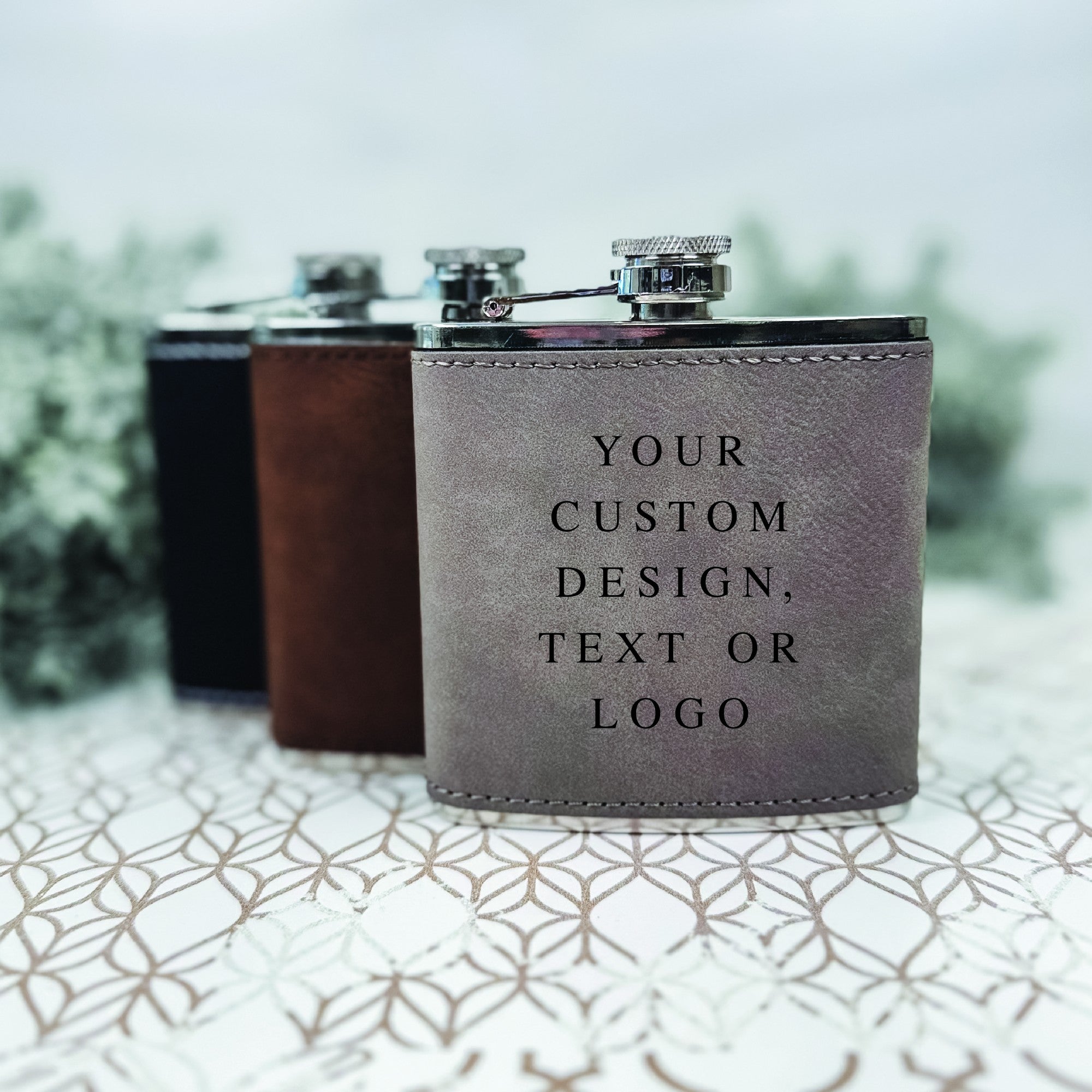 A gray leatherette-wrapped stainless steel hip flask is prominently displayed with a polished silver cap, while brown and black leatherette flasks are softly blurred in the background, serving as versatile personalized gifts for events.