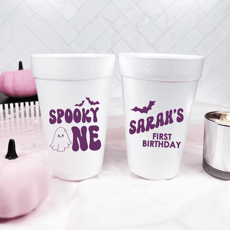 White foam cups with purple text reading "Spooky One" with ghost design and "Sarah's First Birthday" with bat design, displayed with pink Halloween decorations.