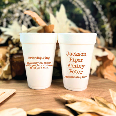 Two white foam cups, personalized with rust-colored text, are displayed on a wooden table adorned with dried autumn leaves, perfect for a fall celebration or gathering.