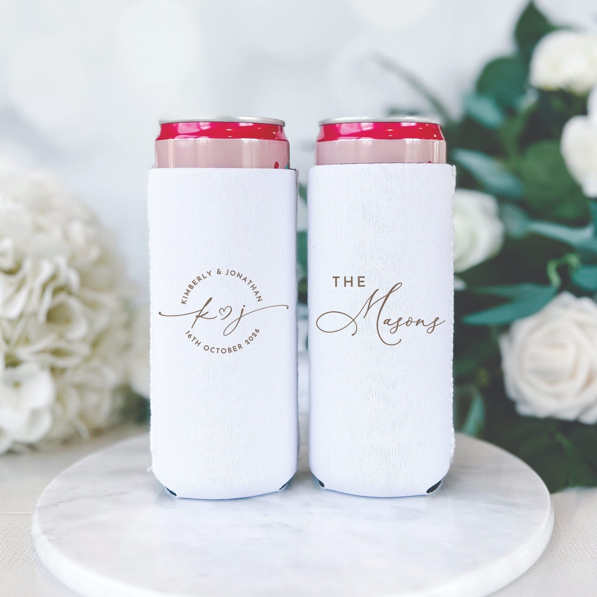 Pair of white slim can coolers with pink checkered trim, one showing a circular logo and the other displaying "THE Mesmer" text in elegant script
