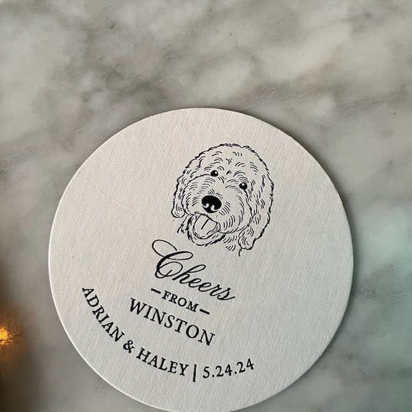 Cream-colored round coaster with black line-drawn dog portrait and personalized text reading "Cheers from Winston" with names and date below.