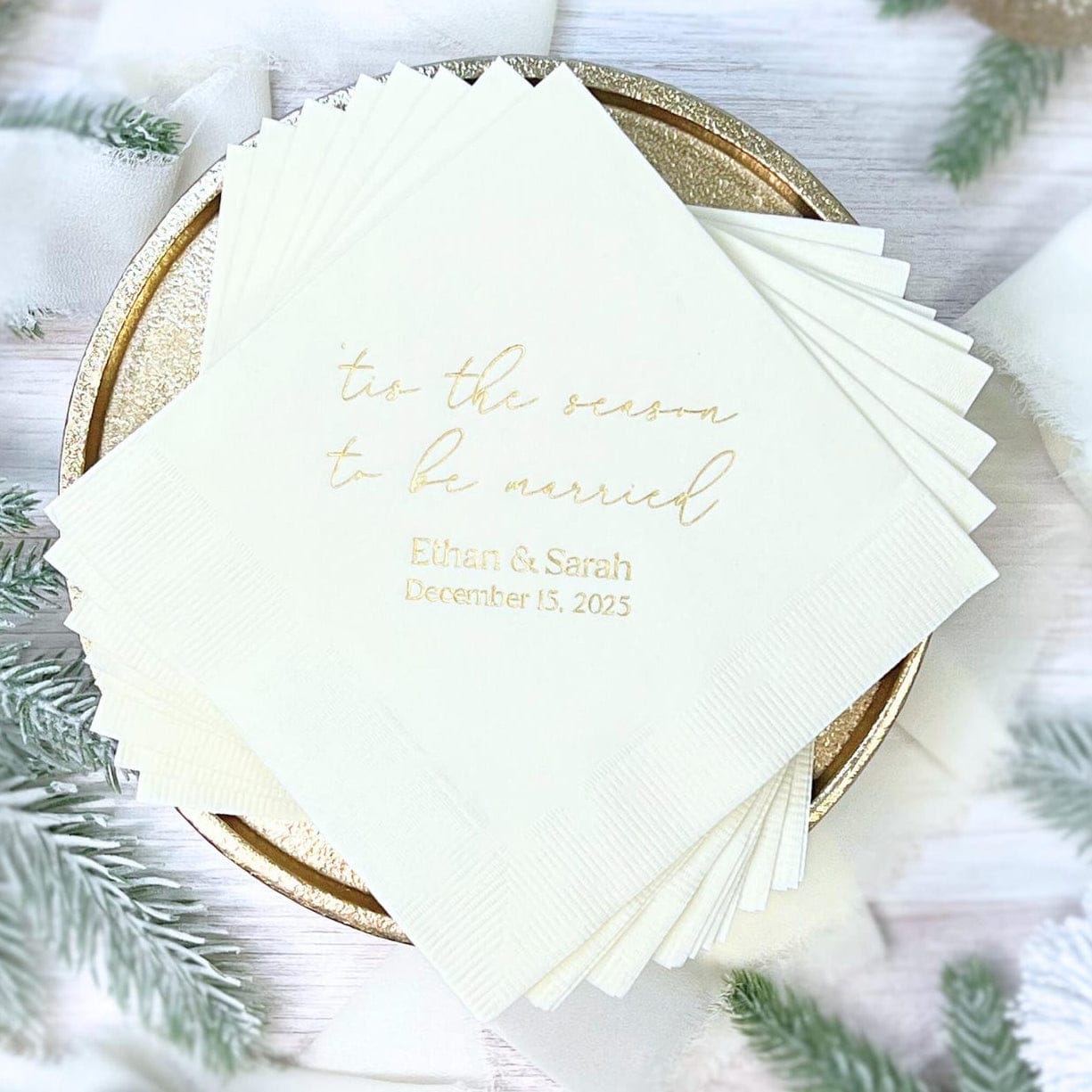 Stack of cream napkins with gold foil text reading "tis the season to be married" and couple names with wedding date, displayed on gold charger plate with greenery.