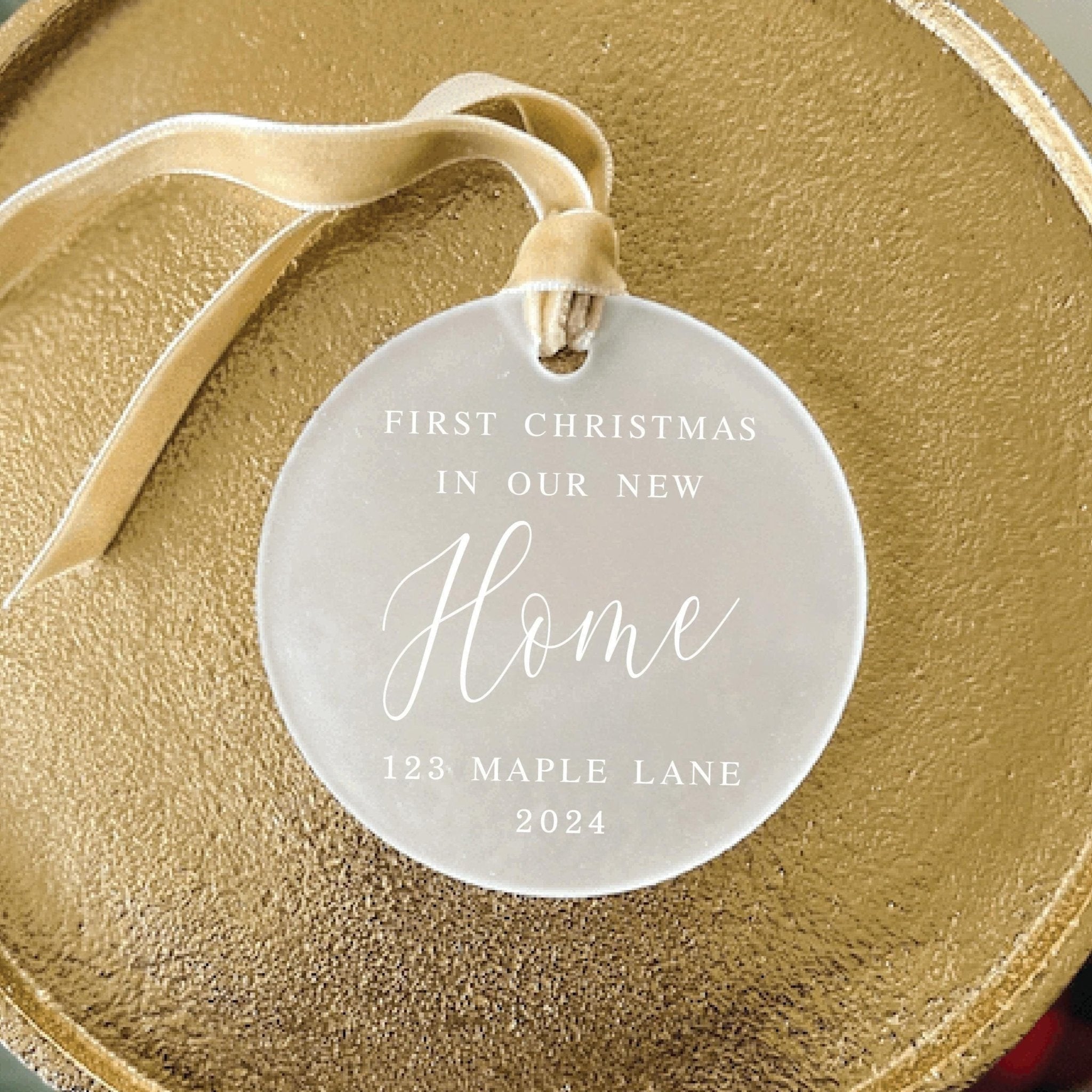 Frosted acrylic round ornament engraved with "First Christmas in Our New Home," address, and year, tied with cream ribbon on gold tray.