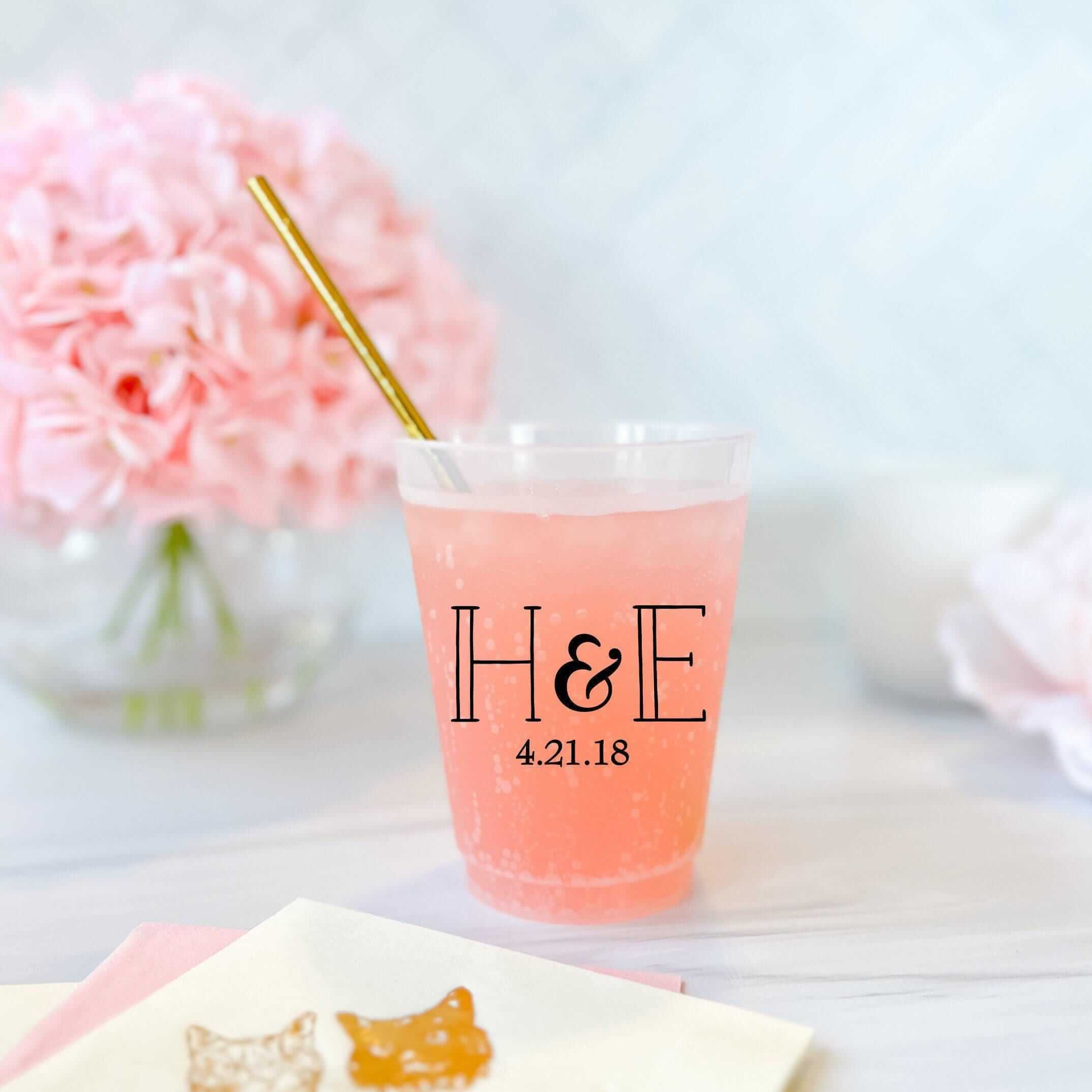 Frosted cup with initials H&E and date 4.21.18, filled with pink beverage and gold straw, surrounded by pink flowers