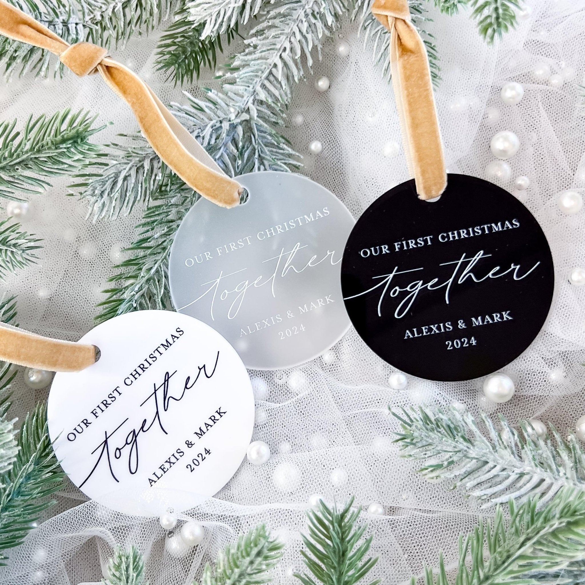 Round acrylic ornaments in white and black with gold ribbon hangers, personalized with "Our First Christmas Together" and couple names, displayed on evergreen branches.